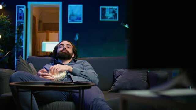 Bored man at home entertaining himself by throwing popcorn in air and catching it with mouth, wasting time. Person slacking off, eating salty snacks and lounging on couch watching TV, camera B