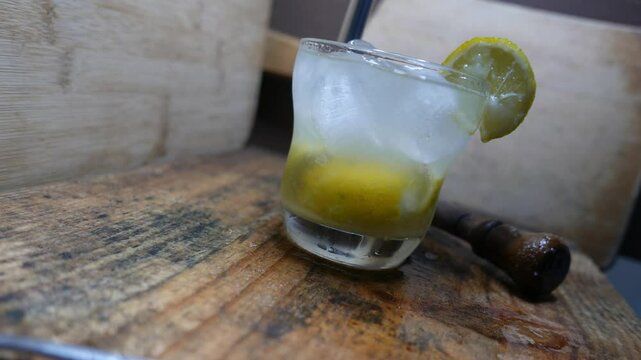A diagonal view of a caipirinha on the table, along with a slow zoom. Someone stirring a glass of caipirinha with a metal drinking straw. Preparing a caipirinha with lemon, cacha&ccedil;a, and sugar.  