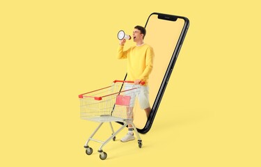 Young man with empty shopping cart shouting into megaphone and big mobile phone on yellow background