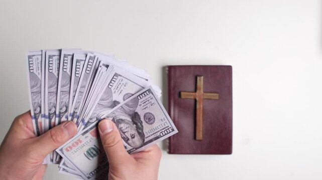 Hands counting one hundred dollar bills near bible with cross: corruption and religion concept