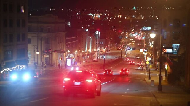 Cars stream through a brightly lit downtown street at night. Headlights and taillights create streaks of color. Buildings glow in the background. Smooth, steady shot.