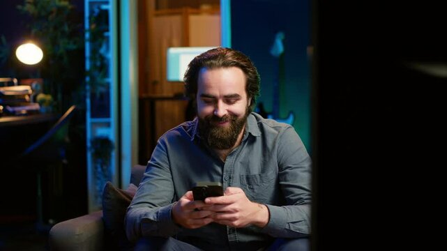 Man sitting on couch at home texting friend on smartphone, inviting him to watch movie on streaming service. Person at home discussing with mate on mobile phone, recommending TV show, camera A