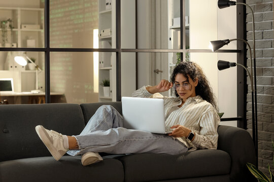Female African-American programmer working with laptop on sofa in office at night