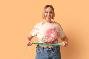 Pretty young woman in loose jeans measuring her waist on beige background. Weight loss concept