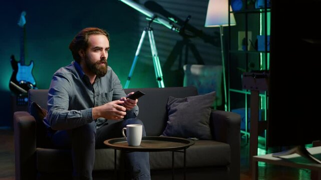 Man appalled by upsetting breaking news story on TV, doing phone call to family member. Person at home scared by troubling events broadcasted on TV, calling friend using smartphone, camera A