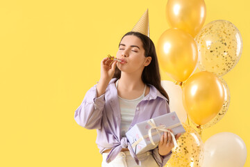 Young woman in party hat with gift and balloons celebrating Birthday on yellow background