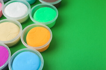 Different colorful modeling clay in plastic jars on green background, closeup. Space for text