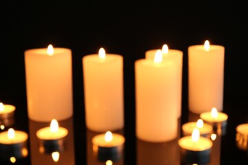 Blurred view of burning candles on mirror surface against black background, closeup