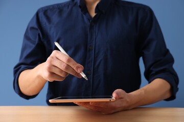 Man with pen working on tablet at desk against blue background, closeup