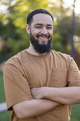 Cheerful African American man poses confidently with arms crossed in a sunny outdoor setting, surrounded by greenery. His friendly demeanor radiates joy, capturing a moment of pure happiness 