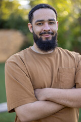 A confident man sporting a stylish beard stands comfortably outdoors, with his arms crossed, presenting a casual and laidback demeanor against a backdrop of lush natural greenery and sunshine