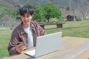 Handsome asian man using laptop computer, mobile phone, working freelance project online, sitting outdoors. Successful business. Asian student studying, learning language, online education concept