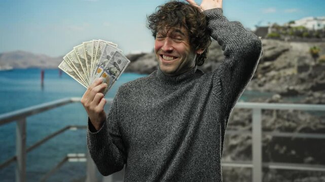 Smiling man holding american dollars by the seaside, reflecting joy and success outdoors on a sunny beach day.