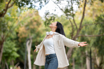 An asian woman is standing with open arms while turning around in the park, breathing in fresh air.