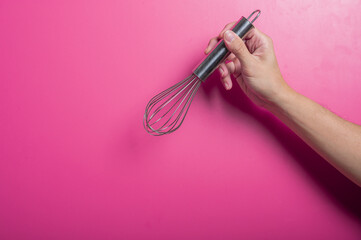 Asian hand holding a stainless steel balloon whisk in front of a pink background. The pose suggests...