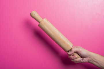 Asian hand gripping a classic wooden rolling pin firmly against a pink background. Used in baking or cooking, the image suggests kitchen preparation, homemade food, or traditional cooking.