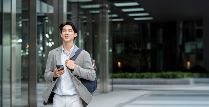 Portrait of young Asian man standing outdoor happy smiling use mobilephone.Handsome young asian man dressed casually walking spending time outdoors at city urban,carrying backpack,using mobile phone - Powered by Adobe