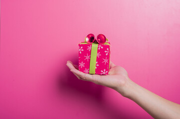 Asian hands holding a small festive red gift box decorated with green ribbon and bells, placed against a pink background. The scene evokes celebration, giving, and holiday joy or surprise.