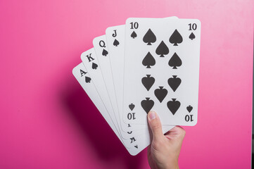 Asian hand holding oversized playing cards showing a royal flush in spades, set against a pink background. The composition suggests winning, gambling, strategy, or luck in a fun visual.