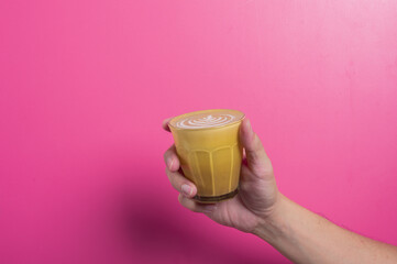 Asian hand holding a glass of hot latte with heart-shaped latte art, set against a pink background....