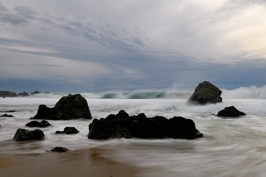 Silhouetted rocks on rugged beach. Long exposure waves paint dreamy surging surf. Beauty of the Big Sur Pacific Coast.