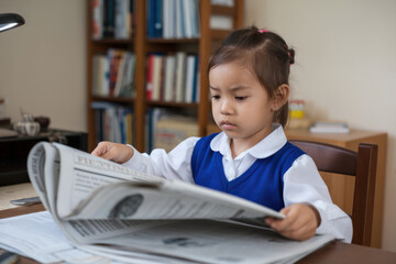 A little girl sitting at a desk, flipping through a financial newspaper with a serious, concentrated expression