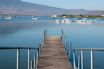 A wooden pier extends over the tranquil waters of Lake Cajititlan in Jalisco, Mexico, with boats and mountains in the background