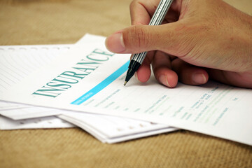 Hand holding pen filling out insurance form on paperwork surface.
