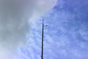A tall metal pole with ladder rungs reaching towards the cloudy blue sky. Minimalist industrial structure, possibly a telecommunications or surveillance tower, captured from a low-angle perspective.

