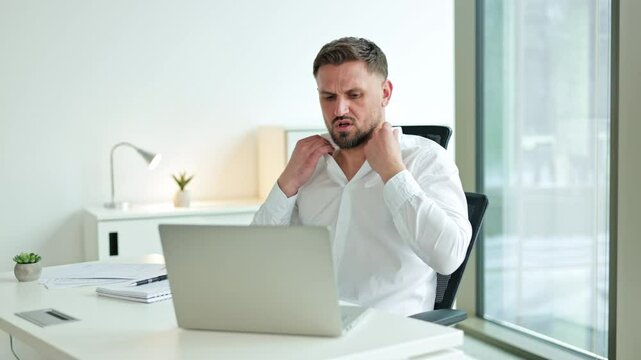 Frustrated businessman sitting at his desk, unbuttoning his shirt and looking at a laptop. The office has a window and a lamp.