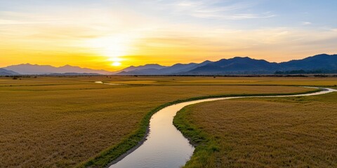 A picturesque landscape featuring a winding river amidst golden fields under a stunning sunset backdrop with mountains in the distance.
