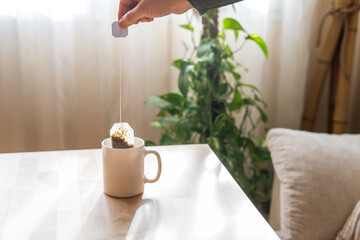 Hand putting a tea bag of chamomile or green tea into a white mug with hot water, on a wooden...