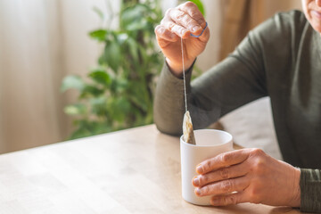 Senior woman enjoying a moment of relaxation, dipping a chamomile and green tea bag into a white mug, sitting at a cozy wooden table in her peaceful home