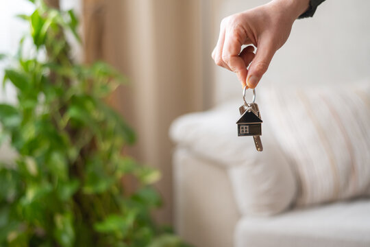 Real estate agent is holding house keys with house shaped keychain in front of blurred sofa in new home, symbolizing home ownership, property purchase, and real estate investment