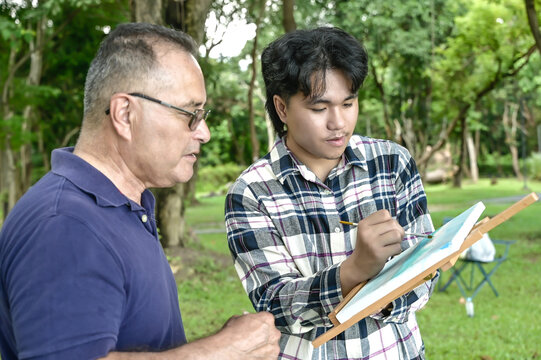 Asian male art student learning art with senior Caucasian teacher in outdoor workshop
