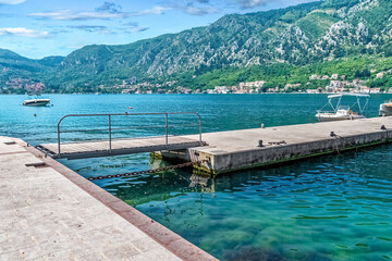 A serene pier extends into the calm waters of Boka Kotorska Bay, with scenic mountains and a peaceful marina in the background. Picturesque view highlights Montenegro natural beauty and maritime charm