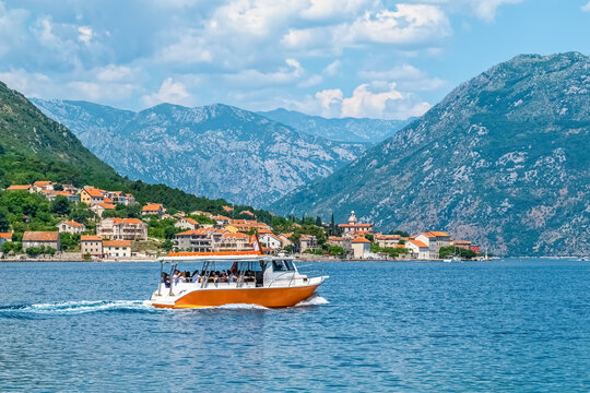 A tour boat cruises through the scenic Bay of Kotor, passing by the charming coastal town of Prčanj, Montenegro, surrounded by majestic mountains. The area is a popular destination for summer travel