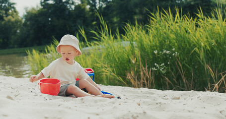 Boy with a hat plays in the sandbox, pouring sand from a bucket, using a strainer, engrossed in...