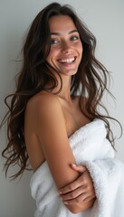 Naklejka premium A beautiful young woman with long, flowing hair smiles warmly while wrapped in a soft, white towel. Her glowing skin and joyful expression radiate confidence and comfort, creating an inviting and