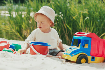 Boy on the beach, pouring sand into a bucket with a shovel, surrounded by toys, his attention fully on the activity near the water.