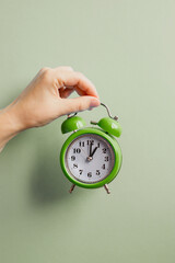 The green alarm clock is held by hand on a light green background. The photo is vertical. Space for...