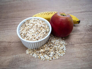 A ceramic bowl filled with raw rolled oats, topped with a ripe banana with brown spots and a red apple. The scene suggests a healthy and natural breakfast.