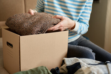 Woman packs donation box with clothes at home. Person places warm sweater into cardboard box....
