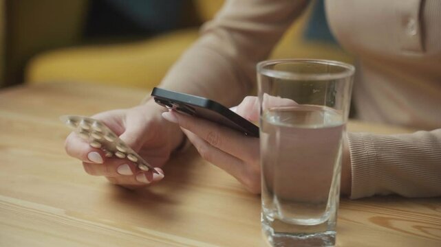 A person examines the instructions for pills while sitting at a table. They hold a smartphone in one hand and a glass of water nearby, focusing on the medication details.