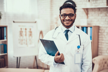 Indian doctor seeing patients in office. Doctor is posing in glasses with clipboard.