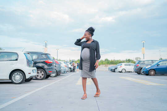 Donna incinta afro con capelli raccolti cammina sorridente in un parcheggio all&rsquo;aperto, indossando un abito grigio aderente e camicia nera, in un&rsquo;atmosfera rilassata con cielo azzurro e auto parcheggi