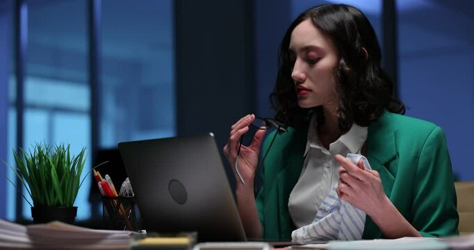Businesswoman wipes glasses with napkin at laptop late at night. Female office employee cleans spectacles in sami dark evening corporate environment