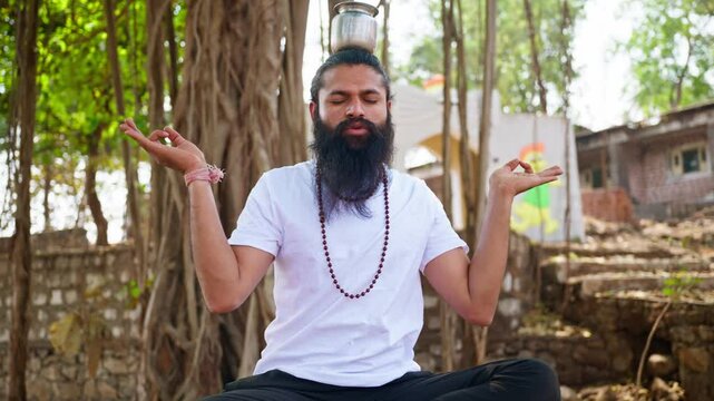 Bearded man meditates balancing a pot on his head outdoor