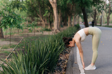 Asian woman stretching before workout outdoors in park, bending forward to touch her toes on paved...
