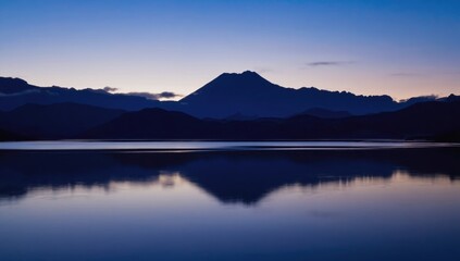 A calm lake reflects the silhouettes of distant mountains during dawn. The sky transitions into soft shades as day breaks, creating a peaceful atmosphere in nature